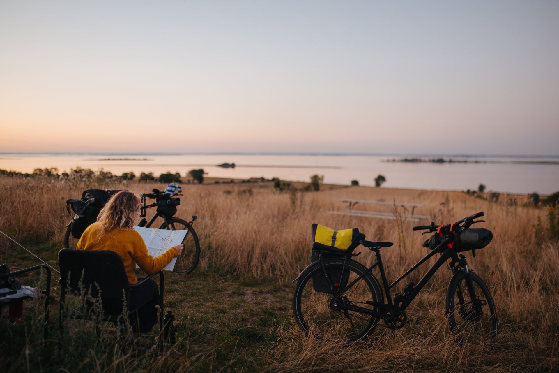 Woman sitting in a camping chair next to her tent and bikes, looking at a map during golden hour, preparing for the next day's cycle