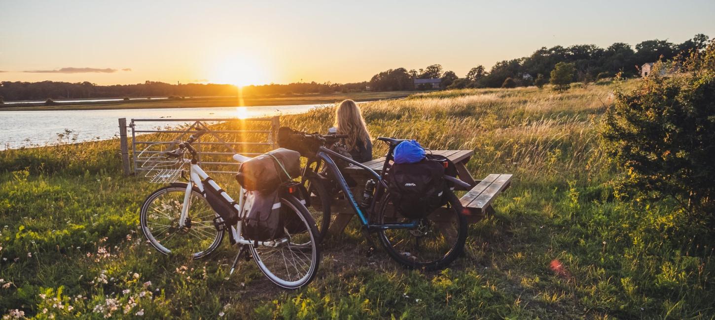 En person sidder i naturen på en bænk med sin cykle i solnedgangen