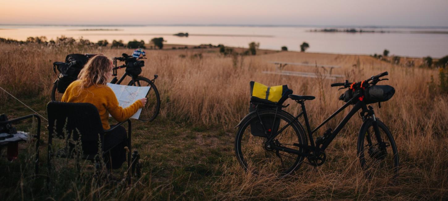 Woman sitting in a camping chair next to her tent and bikes, looking at a map during golden hour, preparing for the next day's cycle